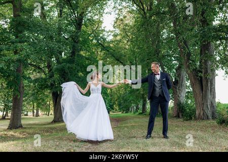 Junger Mann Bräutigam hält Hand der jungen hübschen Frau, die einander anschaut, Braut heben Kante des Kleides. Hochzeit, Tanz. Stockfoto