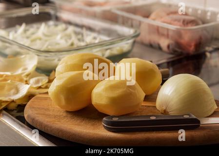 Geschälte Kartoffeln auf einem Schneidebrett und einem Messer, im Hintergrund einige Tabletts mit Zwiebeln und unscharfen Hähnchenschenkeln. Stockfoto
