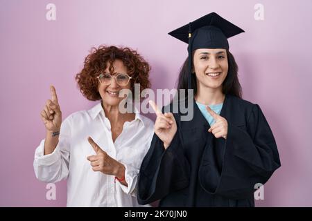 Die hispanische Mutter und Tochter tragen eine Abschlusskappe und eine Zeremonie-Robe und lächeln und schauen auf die Kamera, die mit zwei Händen und Fingern auf den sid zeigt Stockfoto