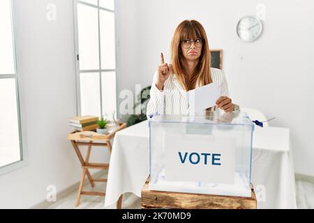 Hispanische Geschäftsfrau beim Abstimmen umhüllt die Wahlurne und zeigt traurig und verärgert auf, zeigt die Richtung mit den Fingern an, unglücklich und niedergedrückt Stockfoto