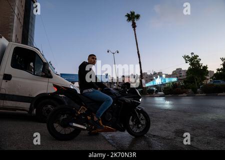 Ramallah, Ramallah und al-Bireh Governorate, Palästina, 12. Juli 2022: Junger, hübscher arabischer Mann in blauer Jeans und schwarzer Jacke sitzt auf einem schwarzen Motorcycl Stockfoto