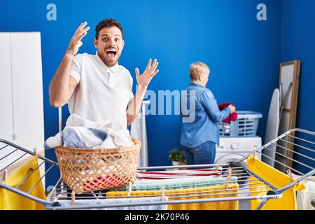 Hispanische Mutter und Sohn hängen Kleider an der Wäscheleine und feiern den Sieg mit glücklichen Lächeln und dem Siegerausdruck mit erhobenen Händen Stockfoto