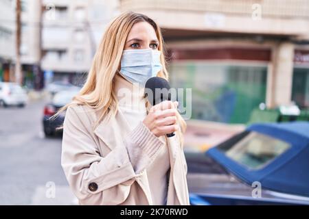 Junge blonde Reporterin trägt eine medizinische Maske und arbeitet auf der Straße Stockfoto