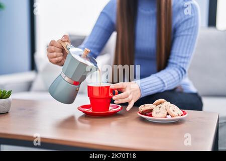 Chinesische Frau, die Kaffee auf die Tasse gießt, sitzt zu Hause auf dem Sofa Stockfoto