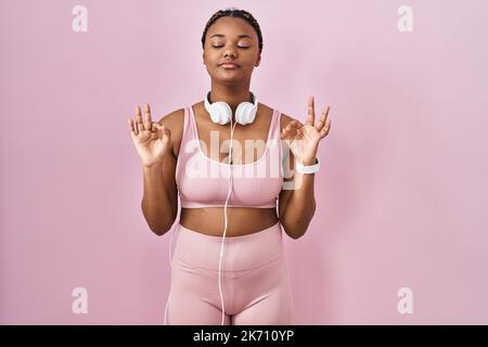afroamerikanische Frau mit Zöpfen, die Sportkleidung und Kopfhörer tragen, entspannt und lächelnd mit geschlossenen Augen und Meditationsgeste mit den Fingern. Yoga Stockfoto