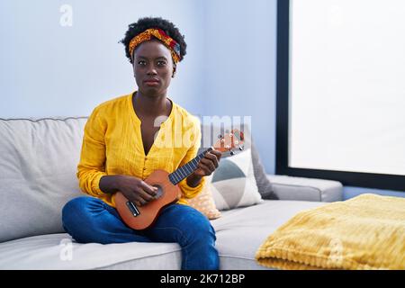 Afrikanische junge Frau, die zu Hause Ukulele spielt, skeptisch und nervös, runzelnd aufgeregt wegen des Problems. Negative Person. Stockfoto