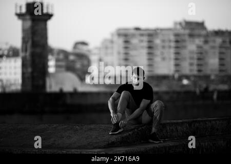 Ein junger Mann sitzt am Meer und schnürt seine Schuhe. Schwarzweiß-Foto. Stockfoto