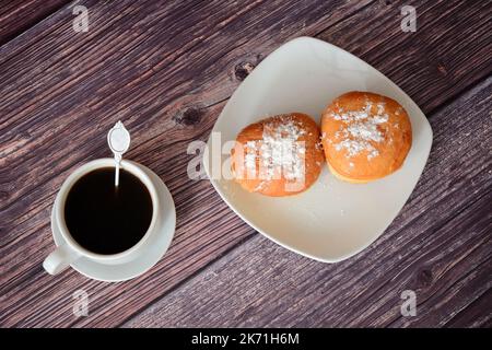 Eine Tasse heißen schwarzen Kaffee und ein Teller mit zwei Donuts mit Puderzucker auf einem Holztisch. Draufsicht, flach liegend. Stockfoto