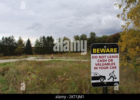 Auf dem Schild steht: Lassen Sie keine Bargeld oder Wertsachen in Ihrem Auto, wenn eine Frau im Hintergrund auf einem Wanderweg im Lakewood Forest Preserve in Illinois unterwegs ist Stockfoto