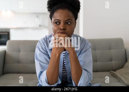 Eine angespannte junge afroamerikanische Frau, die sich über die alltäglichen Probleme im Zusammenhang mit der Krise auslässt, sitzt auf dem Sofa Stockfoto