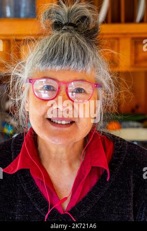 68-jährige Frau chinesischer/italienischer Herkunft mit roter Lesebrille und kabelgebundenen Ohrhörern Stockfoto