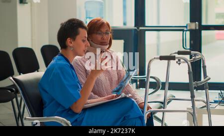 Asiatische Frau mit Halsband beim Betrachten der Röntgenscanergebnisse und Unterstützung durch die Krankenschwester. Assistent, der dem verletzten Patienten, der eine Nackenschaumhalterung trägt, die Diagnose der Radiographie erklärt. Stockfoto