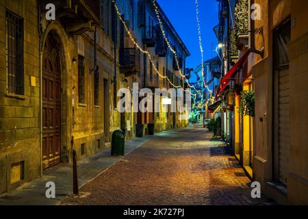 Blick auf die enge Kopfsteinpflasterstraße zwischen alten Gebäuden, die abends in Alba, Piemont, Norditalien, mit Weihnachtslichtern beleuchtet sind. Stockfoto