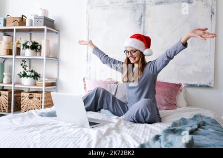 Happy girl in santa Hut grüßt Freunde fröhlich weihnachten Video-Chat auf Laptop. Junge Frau, die in blauem Schlafanzug auf dem Bett liegt, mit geschmücktem Kiefernbaum im häuslichen Interieur mit Kopierraum Stockfoto