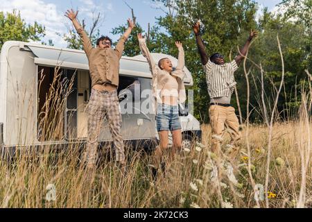 Eine Gruppe von drei Freunden, die mit den Händen in die Luft springen. Millennials haben gemeinsam Spaß vor ihrem Camper auf der Wiese. Sommerzeit. Hochwertige Fotos Stockfoto