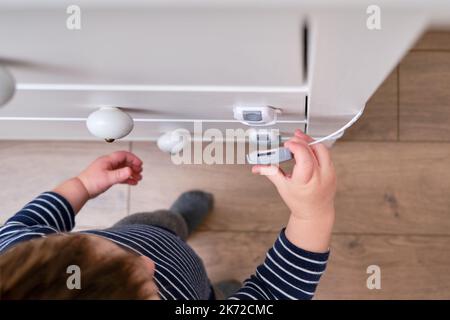 Das Baby öffnet das Kinderschloss in der geschlossenen Schublade des Schranks. Kleinkind Baby mit Hand öffnet Türschloss auf der Brust der Schubladen. Kind im Alter von einem Jahr und drei Stockfoto