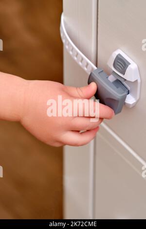 Das Baby öffnet das Kinderschloss in der geschlossenen Schublade des Schranks. Kleinkind Baby mit Hand öffnet Türschloss auf der Brust der Schubladen. Kind im Alter von einem Jahr und drei Stockfoto