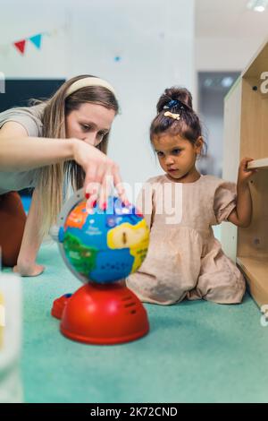 Junge Lehrerin mit langen Haaren, die den Globus spinnen und einem kleinen dunkelhaarigen Mädchen die Dinge erklären, vertikale Aufnahme im Kindergarten. Hochwertige Fotos Stockfoto