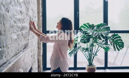 Kreative junge Frau schmückt ihre Wohnung im Loft-Stil, indem sie Platz auf einer Ziegelwand für ein schönes Bild und eine Markierung mit Bleistift wählt. Kreativität und Raumkonzept. Stockfoto