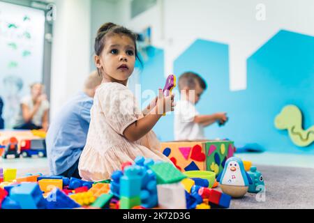 Entzückendes kleines Mädchen, das in den Spielzeugen sitzt und mit großem Interesse irgendwo im Kindergarten sucht. Hochwertige Fotos Stockfoto