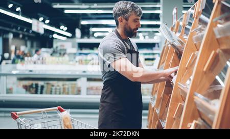 Ein vielbeschäftigter männlicher Verkäufer legt in der Bäckerei im Lebensmittelgeschäft Brot in die Regale, ein bärtiger Mann trägt eine Schürze. Verkauf von Produkten, Beruf und Handelskonzept. Stockfoto