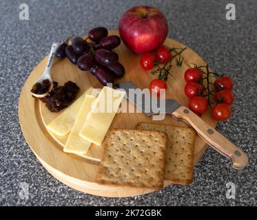 Ein Käseschneidebrett und ein Käsemesser mit geschnittenem Käse, Kirschtomaten, einem roten Apfel, roten Trauben, Crackern, Chutney Picke und ein Esslöffel, auf Stockfoto