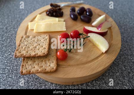 Ein Käsebrett mit Käse, Kirschtomaten, roten Apfel, roten Trauben, Crackers Chutney Pickle und einem Esslöffel, auf einer Küche wo Stockfoto