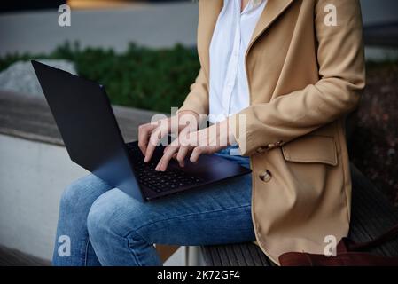 Frau, die aus der Ferne arbeitet, auf der Bank an der Stadtstraße sitzt und einen Laptop benutzt. Freiberufliche Mitarbeiterin arbeitet online und tippt auf der Laptop-Tastatur Stockfoto