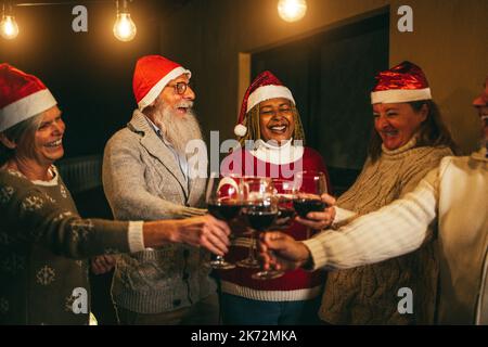 Verschiedene ältere Menschen, die am Heiligabend mit Wein jubeln und Weihnachtsmannmützen tragen - weicher Fokus auf afrikanisches Frauengesicht Stockfoto