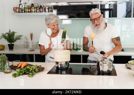 Glückliches Seniorenpaar, das Spaß hat, zu Hause zu tanzen und zu kochen - konzentrieren Sie sich auf die Hand der Frau, die Gurke hält Stockfoto