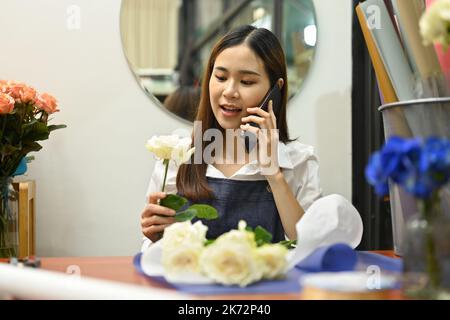 Eine hübsche junge Frau, die mit dem Handy spricht und Bestellungen von Kunden entgegennimmt, während sie in ihrem Blumengeschäft sitzt Stockfoto