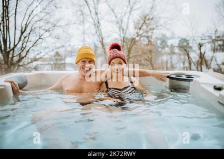 Das ältere Paar mit kitschiger Kappe genießt an kalten Wintertagen gemeinsam die Badewanne im Freien auf der Terrasse. Stockfoto