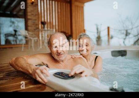 Ältere Paare genießen gemeinsam eine Badewanne im Freien auf ihrer Terrasse während des kalten Wintertages. Stockfoto