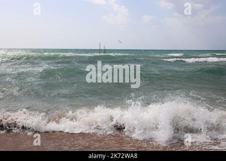 Wellen brechen an der sandigen Küste mit Spritzern. Die Stäbe, die die Fischernetze halten, ragen aus dem Wasser. Eine Möwe fliegt in der Ferne. Stockfoto