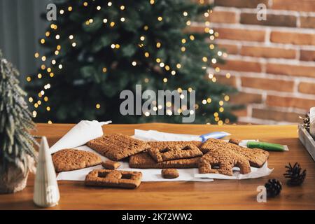 Hausgemachte Lebkuchen Haus Bausteine mit Glasur auf Tisch mit Dekorationen, Laternen im Wohnzimmer mit defokussierten Silvester Lichter, Weihnachtstanne dekoriert. Urlaubsstimmung für Kinder Stockfoto