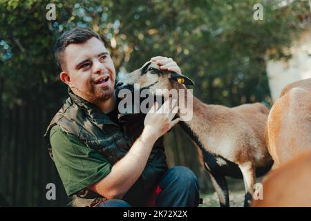Hausmeister mit Down-Syndrom kümmert sich um Tiere im Zoo, streichelte Ziege. Konzept der Integration von Menschen mit Behinderungen in die Gesellschaft. Stockfoto