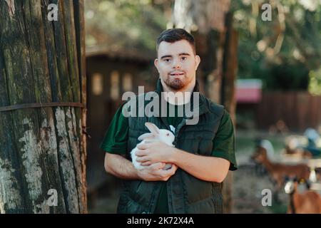 Hausmeister mit Down-Syndrom kümmert sich um Tiere im Zoo, streicheln Kaninchen. Konzept der Integration von Menschen mit Behinderungen in die Gesellschaft. Stockfoto