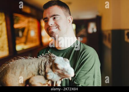 Porträt eines Hauspflegers mit Down-Syndrom, der sich im Zoo um Tiere kümmert, streichelt Leguan. Konzept der Integration von Menschen mit Behinderungen in die Gesellschaft. Stockfoto