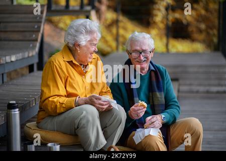 Glückliches Seniorenpaar in Herbstkleidung, das nach dem Spaziergang in der Nähe des Sees Picknick macht. Stockfoto