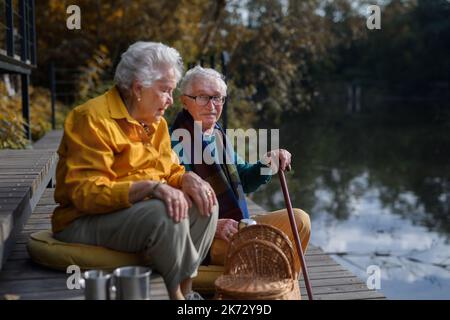 Glückliches Seniorenpaar in Herbstkleidung, das nach dem Spaziergang in der Nähe des Sees Pause gemacht hat. Stockfoto