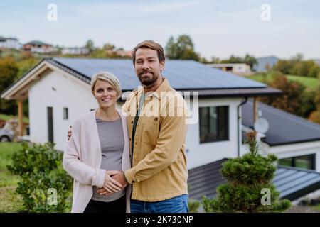 Glücklicher Mann mit seiner Schwangeren Frau, der vor ihrem neuen Haus mit Photovoltaik-Sonnenkollektoren steht. Stockfoto