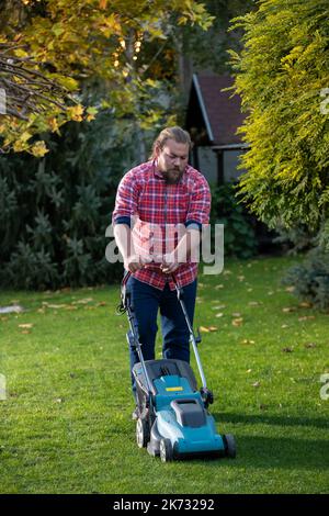 Schöner Mann mit Bart, der im Herbst Rasenmäher schiebt und Gras in seinem Hinterhof mäht Stockfoto