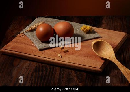 Zwei braune Hühnereier auf Quappe Serviette über Schneidebrett und Holzlöffel auf dunklem Tisch und warmen rustikalen Hintergrund Stockfoto