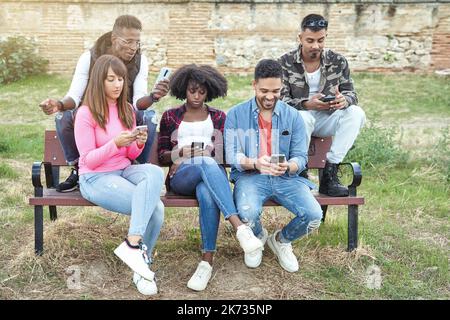 Eine Gruppe von multiethnischen Freunden, die ihr Mobiltelefon benutzen, während sie auf einer Bank sitzen. Technologie und Freundschaftskonzept. Stockfoto