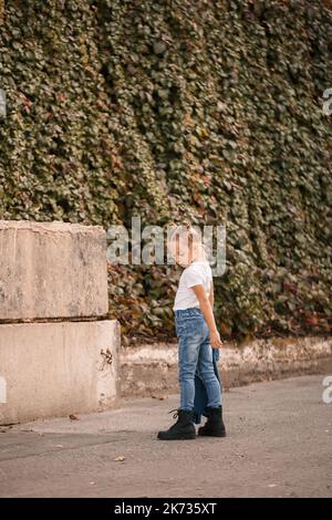 Stylisches blondes kleines Mädchen in Jeans und einem weißen T-Shirt geht die Straße hinunter. Mädchen 7 Jahre alt kleines Modell, schönes Kind Stockfoto