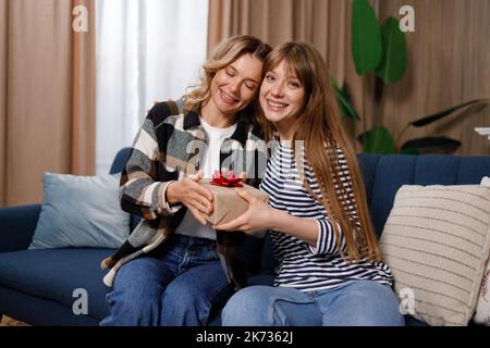 Die Erwachsene Frau überrascht ihre Mutter im mittleren Alter unerwartet und gibt ihr eine Geschenkbox, während sie auf dem Sofa in der Wohnung sitzt Stockfoto
