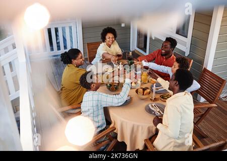 Blick aus dem hohen Winkel auf große afroamerikanische Familie klirrende Gläser über dem Esstisch im Freien in gemütlicher Umgebung mit Lichterketten Stockfoto