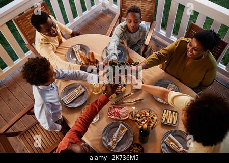 Blick aus dem hohen Winkel auf große afroamerikanische Familie klirrende Gläser über dem Esstisch im Freien in gemütlicher Abendumgebung Stockfoto