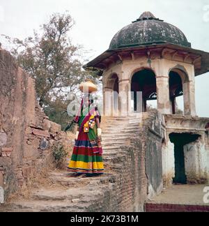 Vintage 1961 Farbfoto zeigt ein junges indisches Mädchen, das gerade Wasser aus einem Brunnen gesammelt hat und das Wasser in einer Pfanne auf dem Kopf trägt, einige Stufen hinunter. Stockfoto