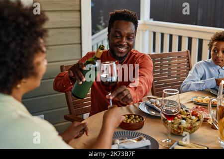 Porträt eines lächelnden schwarzen Mannes, der Wein in die Gläser gießt, während er die Abendparty im Freien genießt Stockfoto